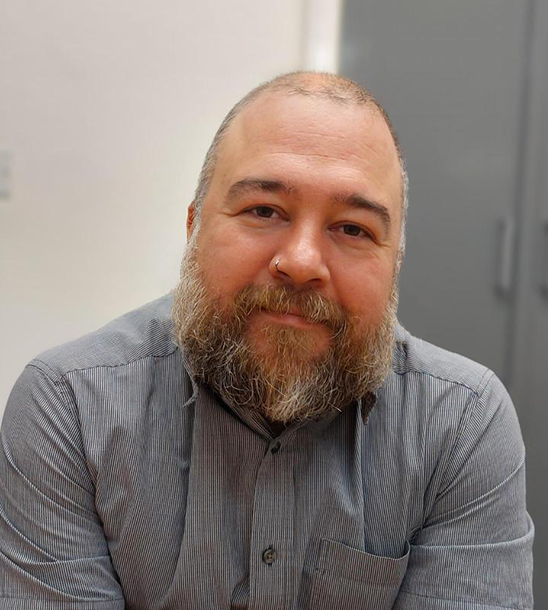 Headshot of Malcolm - wearing a grey checked shirt, with shaved head and unkempt beard.