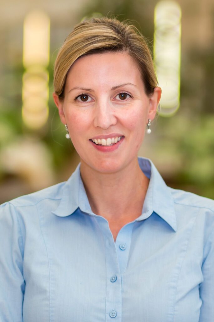 Headshot of Sarah with blonde hair and pearl earrings wearing a light blue blouse looking to the camera in front of a blurred background