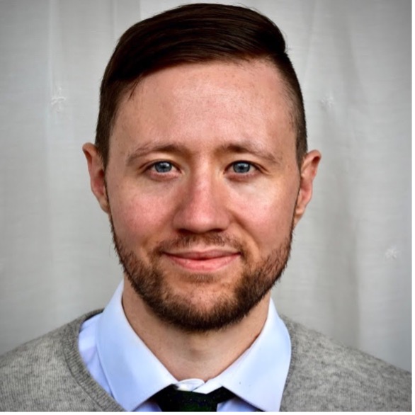 Headshot of Thomas wearing a blue shirt and grey sweater in front of a grey background. He has a short beard and side-parted hair.
